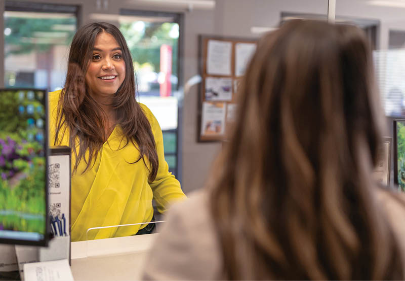 A beautiful Latina woman smiles while speaking with a friendly customer service representative at a reception desk outfitted with an acrylic glass sneeze guard.