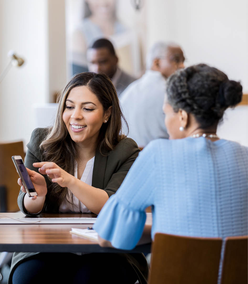 Mid adult female bank employee discusses a mobile banking app's features with a mature female client.