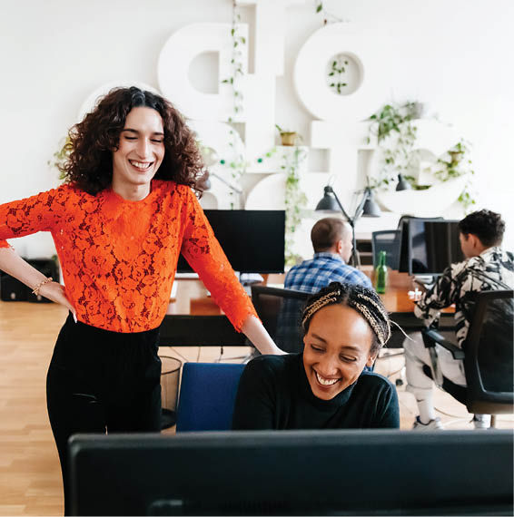 Two office workers smiling. one working on computer while other watches